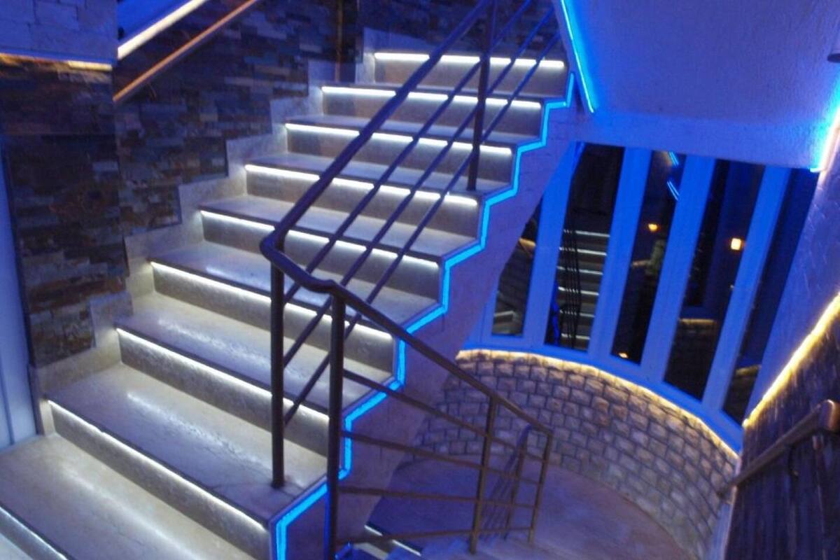 A staircase with illuminated handrails and steps, leading to a landing with a stone wall and windows.