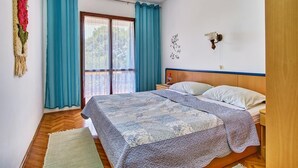 A bedroom with a bed, a nightstand, a wall-mounted lamp, and a view through a window with curtains. - Apartments Pero (Mali Losinj)