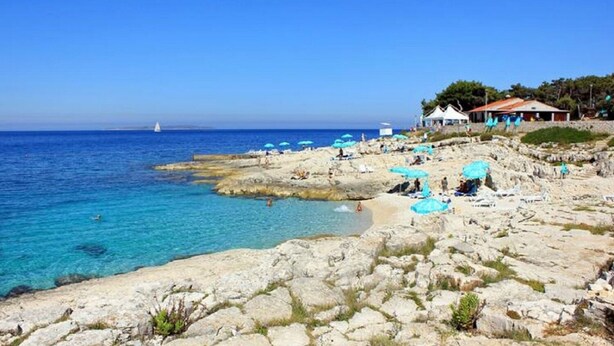 A rocky beach with clear blue water, a few umbrellas, and a building with a red roof. - Apartments Pero (Mali Losinj)