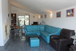 Ein Wohnzimmer mit einem blauen Ecksofa, einem braunen Ledersessel, einem Glastisch und einem Essbereich mit Stühlen und einem Tisch.