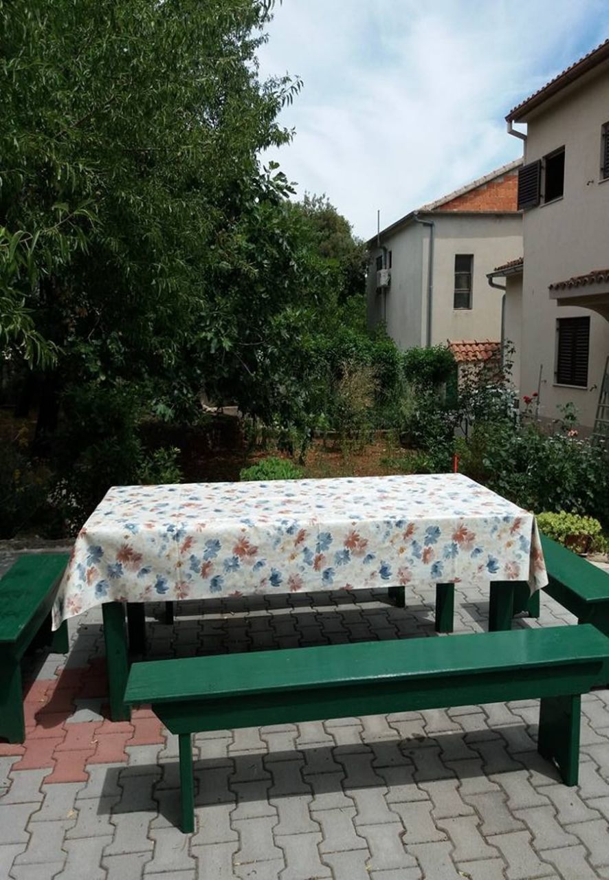 A picnic table with a floral tablecloth, green benches, and a paved patio area with a house and trees in the background.