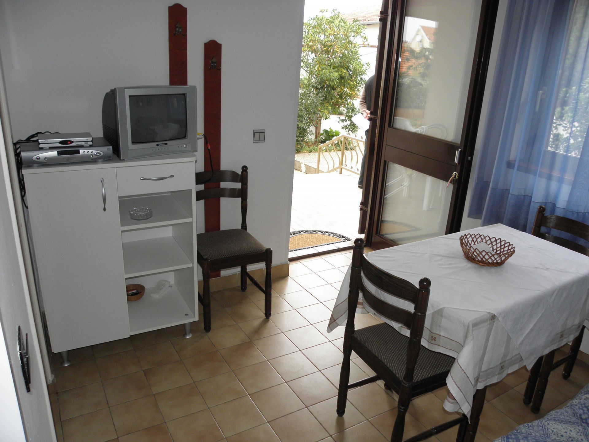 A small room with a television, a cabinet, a chair, a table with a white tablecloth, and a window with curtains.
