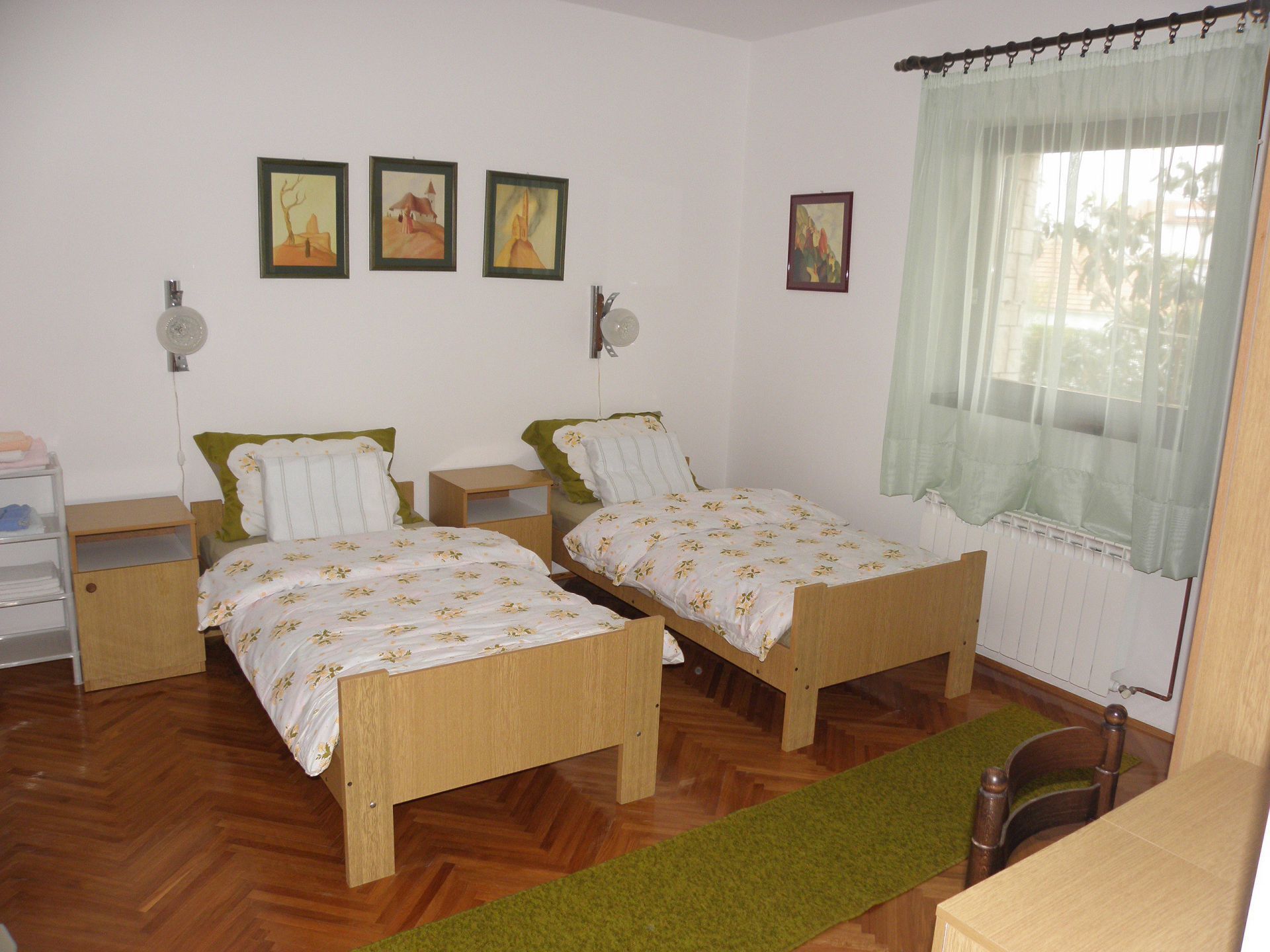 A room with two wooden beds, a desk, and a chair. There are three framed pictures on the wall.