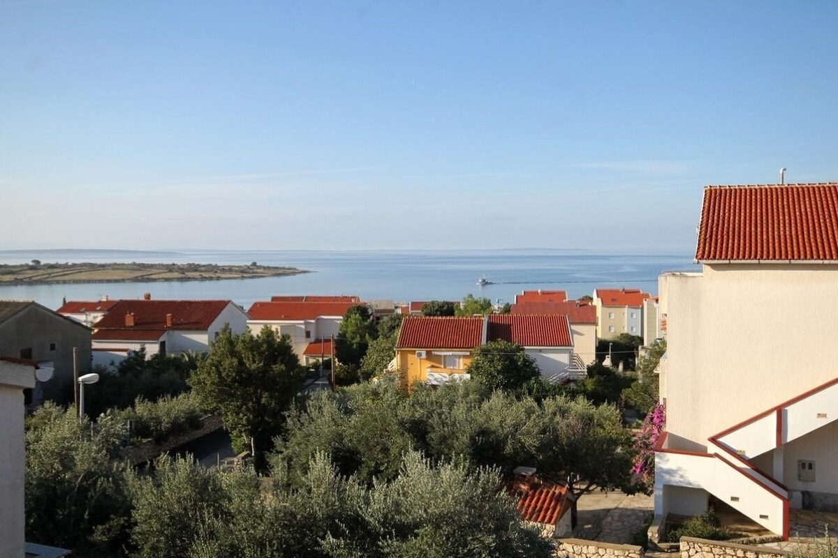 A coastal residential area with houses featuring red-tiled roofs, a clear view of the sea, and a stone pathway leading to a building with a white facade.