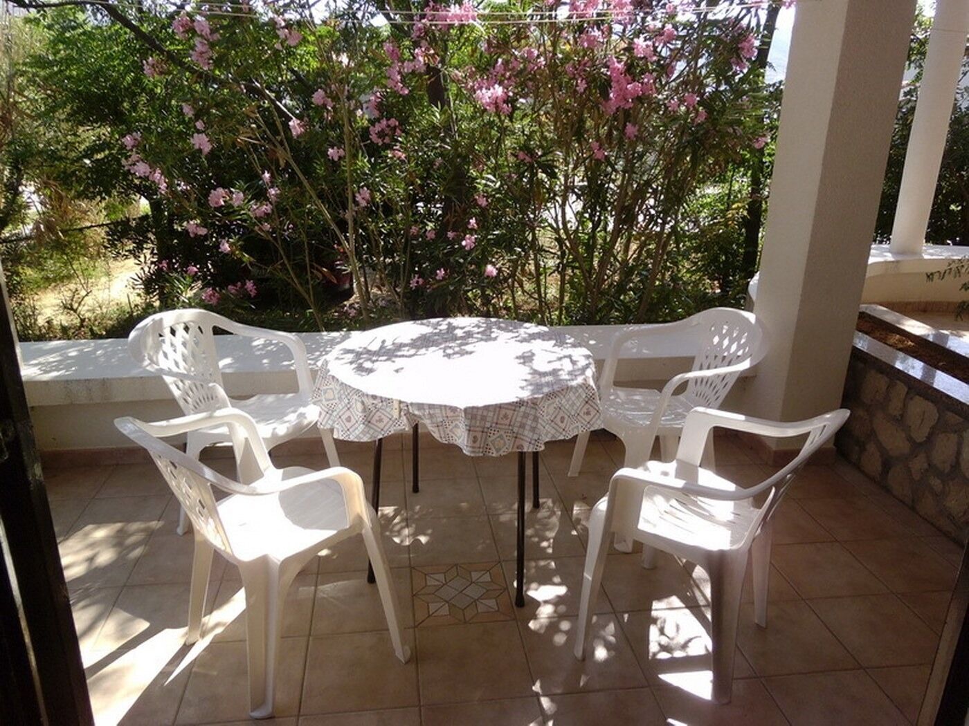 Une table ronde entourée de quatre chaises en plastique blanc, recouverte d’une nappe blanche, est placée sur une terrasse carrelée. Un mur de pierre et des plantes en fleurs se trouvent en arrière-plan.