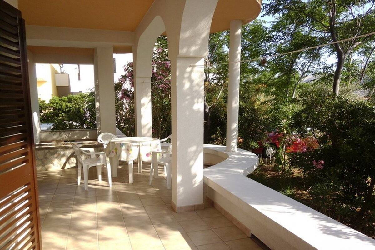 A covered outdoor patio with a round table and chairs, surrounded by white columns and a garden with various plants and flowers.