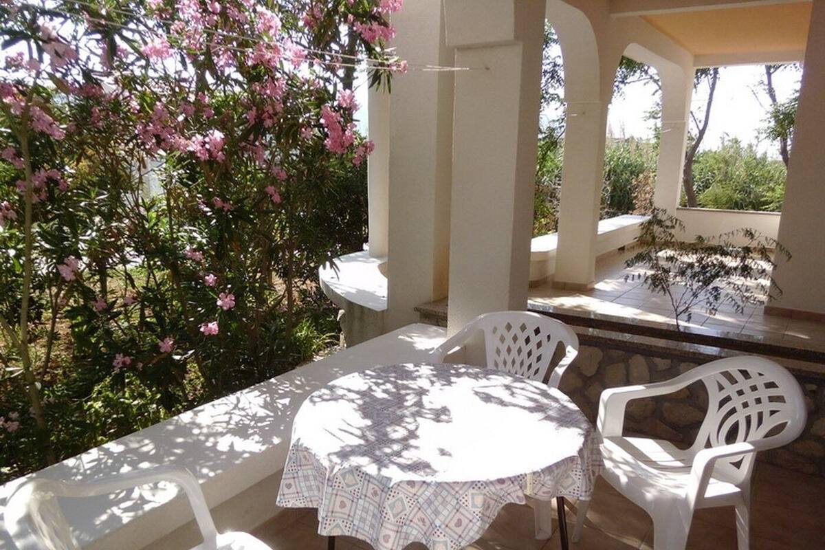 An outdoor patio with a round table and white plastic chairs, surrounded by white pillars and covered by a pergola.