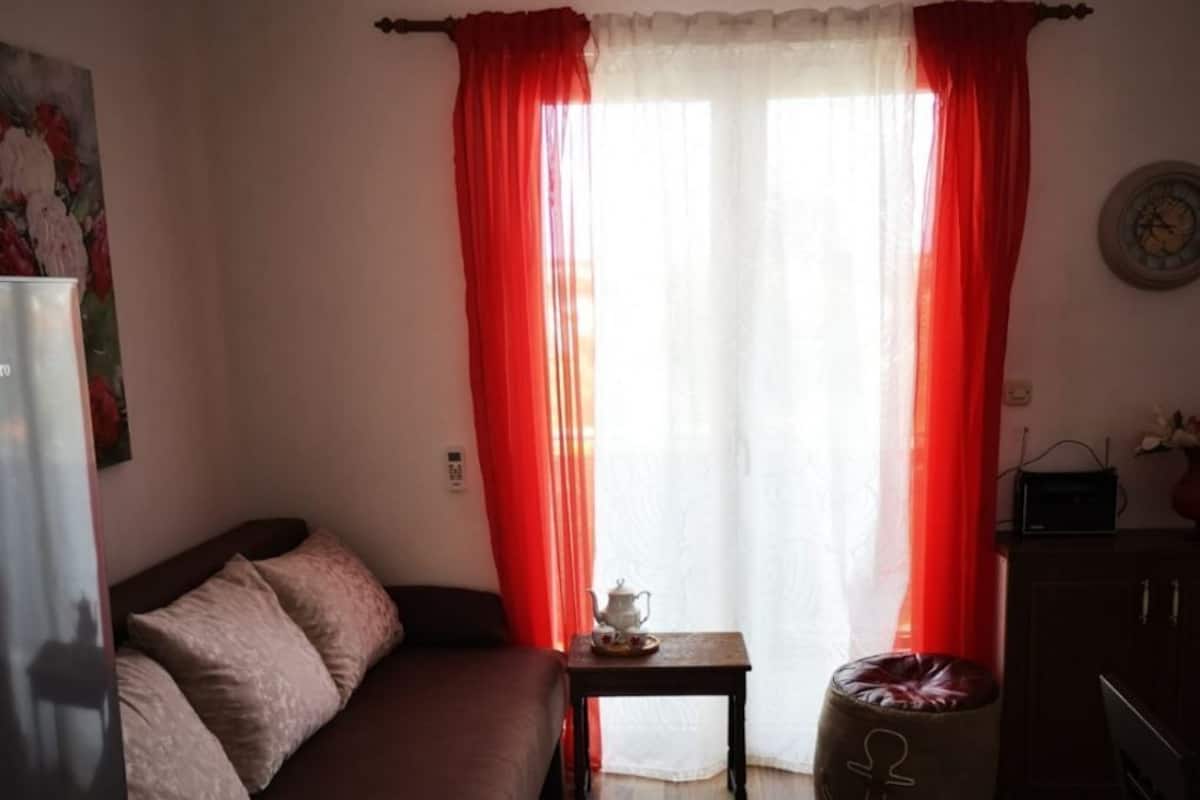 Une petite pièce chaleureuse, avec un plancher en bois, un canapé marron, une petite table sur laquelle se trouve une théière, et un rideau rouge.