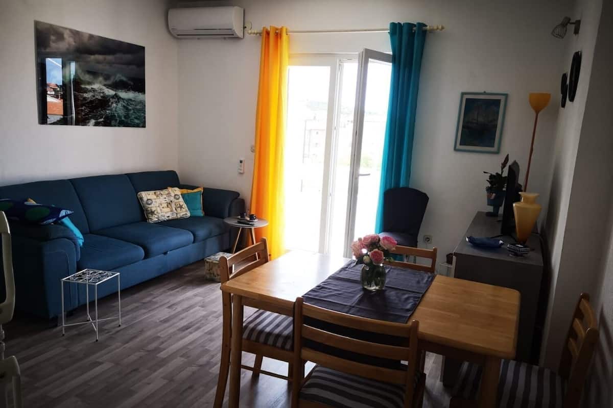 Un salon avec un canapé bleu, une table à manger en bois avec des chaises, une petite table d’appoint et une fenêtre avec des rideaux.