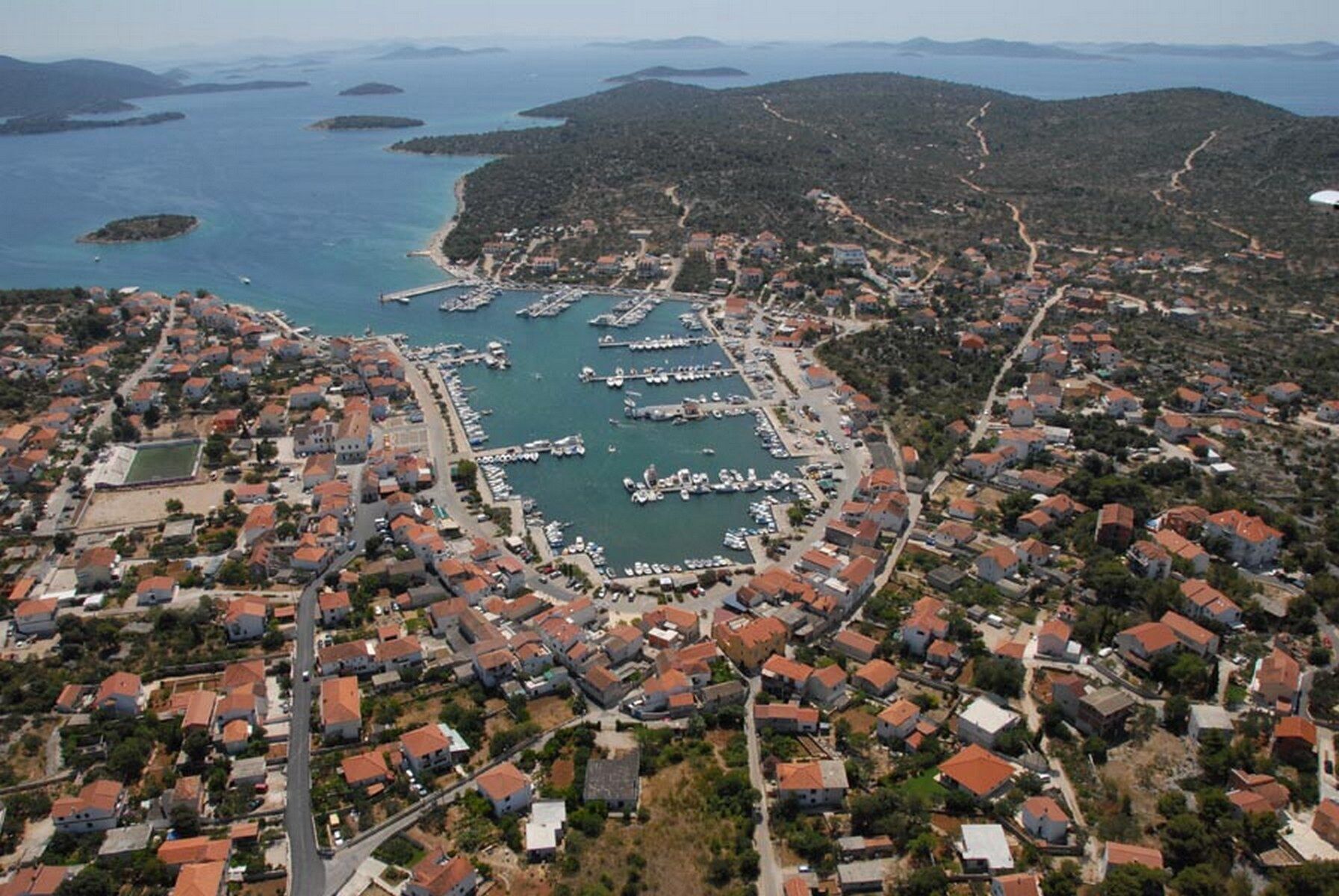 An aerial view of a coastal town with a marina, residential buildings, and surrounding hills.