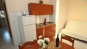 Een kleine keuken met een koelkast, spoelbak en houten kastjes. Op de voorgrond staat een eettafel met stoelen en een bloemenvaas.
