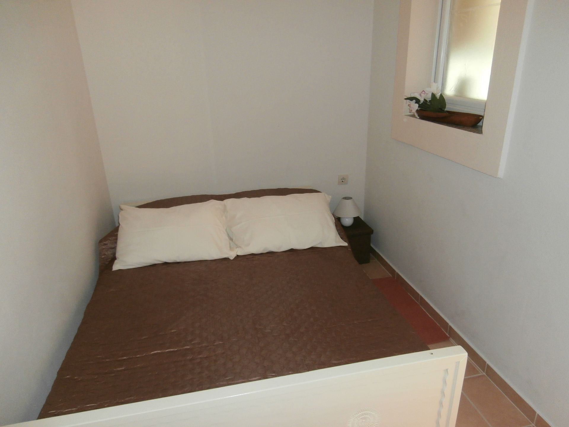 Ein kleines, schmales Zimmer mit einem Einzelbett, einem Nachttisch, einer Lampe und einem Fenster mit Blick nach draußen.