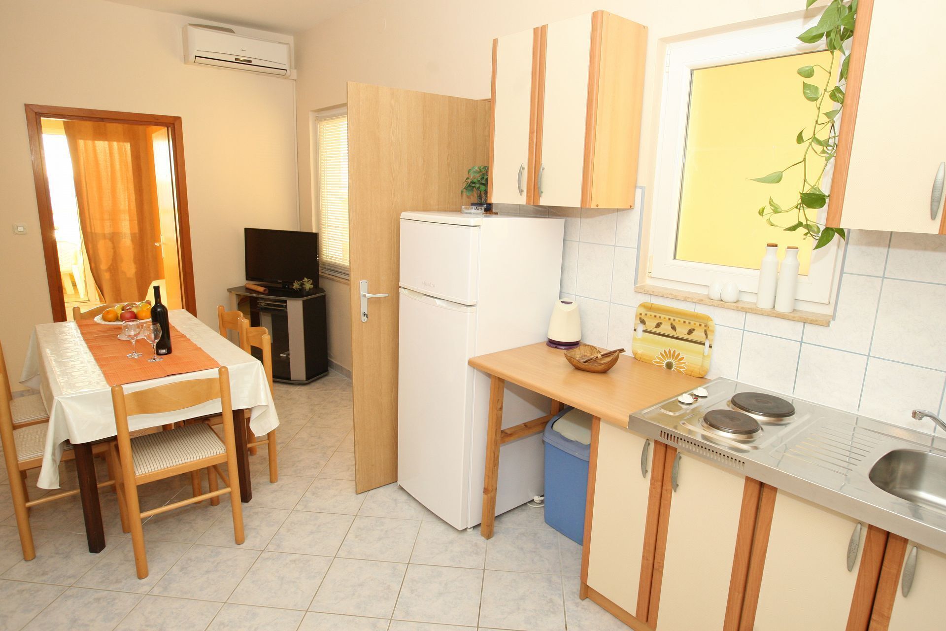 A compact kitchen with a dining area, a small refrigerator, a sink, and a window with a plant.