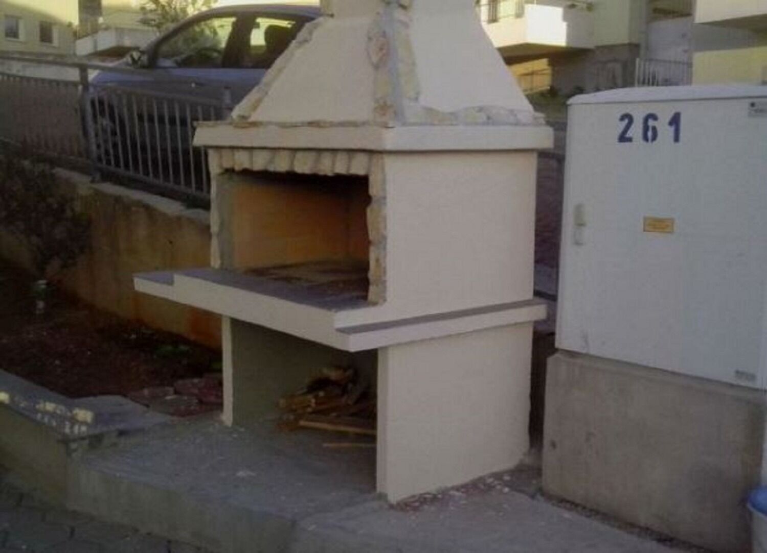 A small, white outdoor structure with a chimney, possibly a wood stove or fireplace, next to a utility box marked with the number 261.