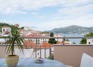A balcony with a potted plant and a table, overlooking a coastal town with red-roofed buildings and a view of the sea and mountains. - Apartment Slavica (Okrug)