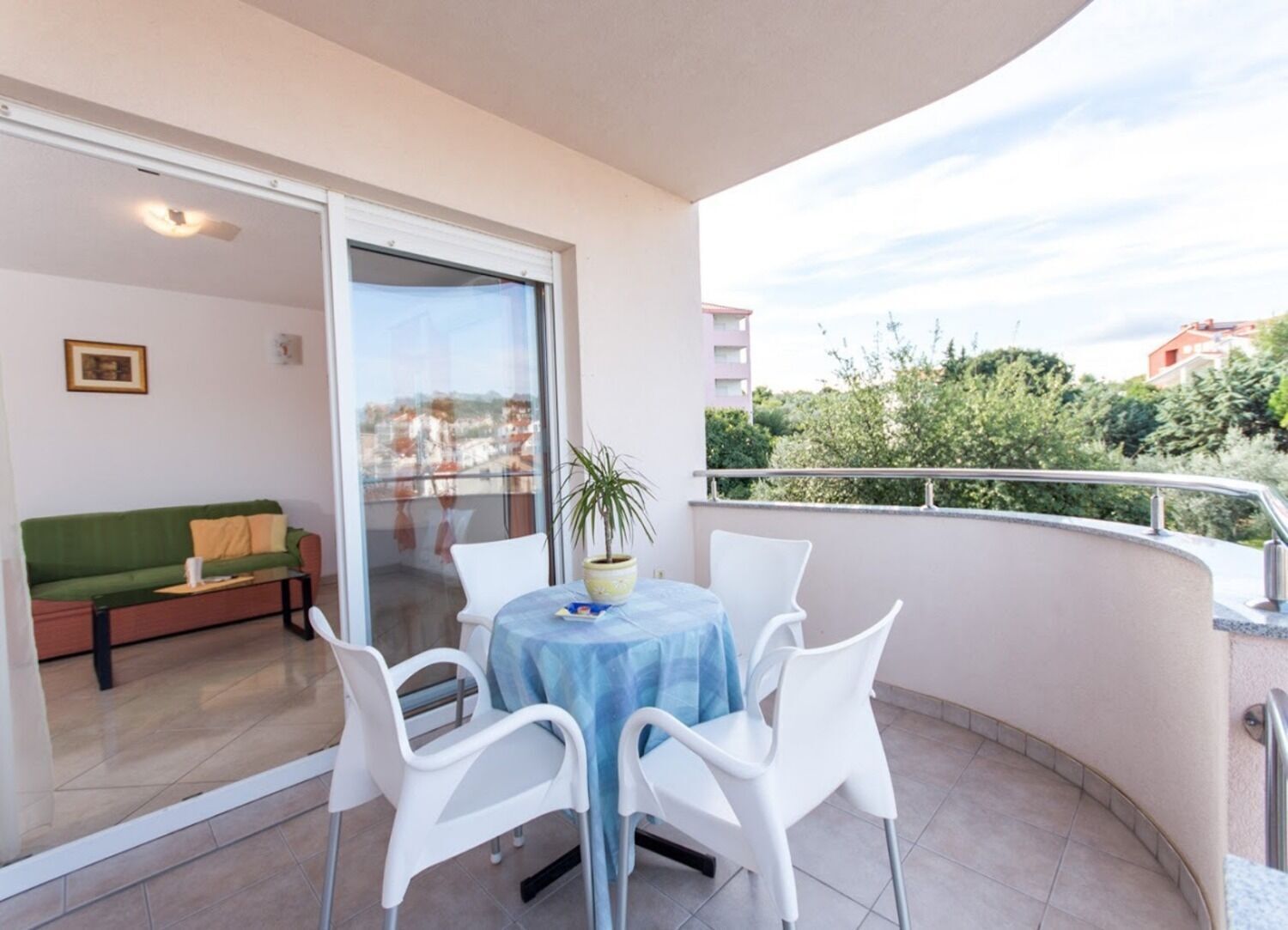 Ein Balkon mit Tisch und Stühlen, einem Topf mit einer Pflanze und einer Schiebetür zu einem Wohnbereich mit einer Couch und Kissen.