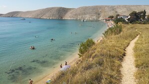 A coastal beach with clear waters, a sandy shore, and a path leading to a row of houses. - Apartments Ana (Pag)