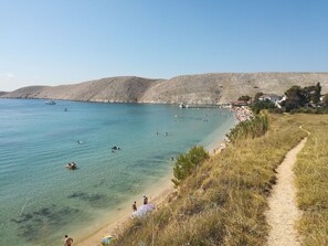 Una playa costera con aguas transparentes, una orilla arenosa y un sendero que conduce a una hilera de casas.