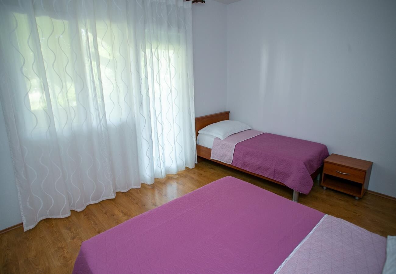 A room with two beds, a wooden floor, a white wall, and a window with sheer curtains.