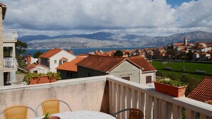 Ein Balkon mit Korbstühlen und einem Tisch, mit Blick auf eine Küstenstadt mit roten Dächern und einem Gebirgszug im Hintergrund.