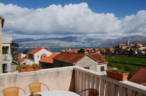 Un balcón con sillas de mimbre y una mesa, con vista a un pueblo costero con edificios de techo rojo y una montaña al fondo.