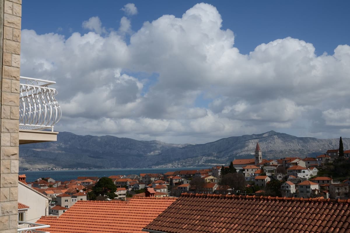 A coastal town with terracotta-roofed buildings, a church with a red spire, and mountains in the background.