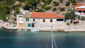 A sailboat docked near a stone building with a red roof, surrounded by calm water and greenery. - House Linker (Vis)