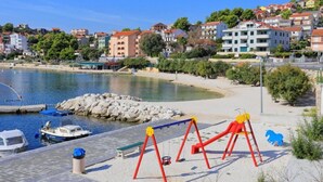 Una zona costera con una playa arenosa, un parque infantil con columpios rojos y un puerto deportivo con barcos.