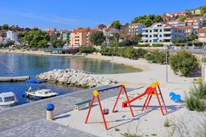 A coastal area with a sandy beach, a playground with red swings, and a marina with boats. - Apartments Pero (Marina)
