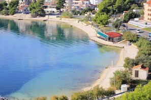 A coastal area with a clear blue waterfront, a small building with a red roof, and residential buildings in the background. - Apartments Pero (Marina)
