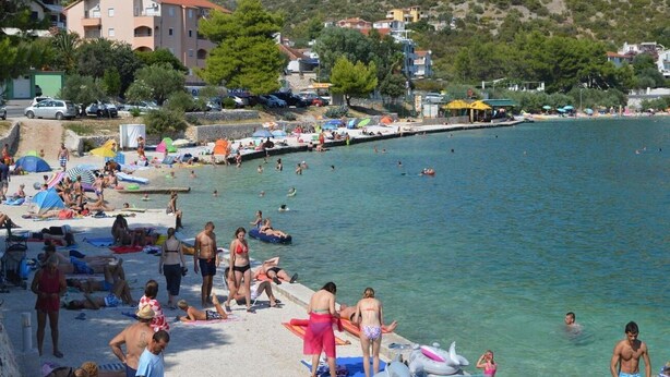 Una playa con gente tomando el sol y nadando, un muelle que se adentra en el agua, y edificios al fondo.