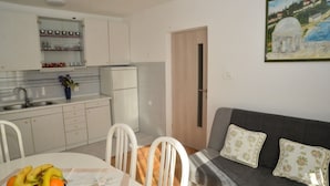 A compact kitchen with white cabinets, a sink, and a small refrigerator. There is a dining table with chairs and a fruit bowl. A gray sofa with floral cushions is against the wall. - Apartment Miki (Zadar)