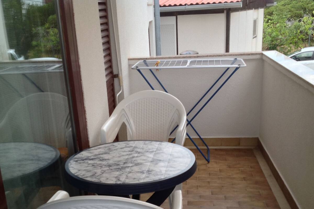 A small balcony with a round table and two chairs, a drying rack, and a view of a building with a tiled roof.