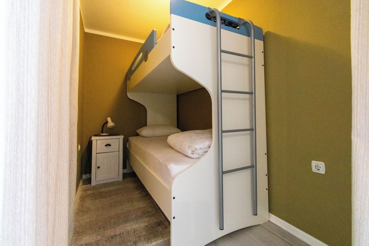A bunk bed with a ladder, a bedside table with a lamp, and a power outlet on the wall.
