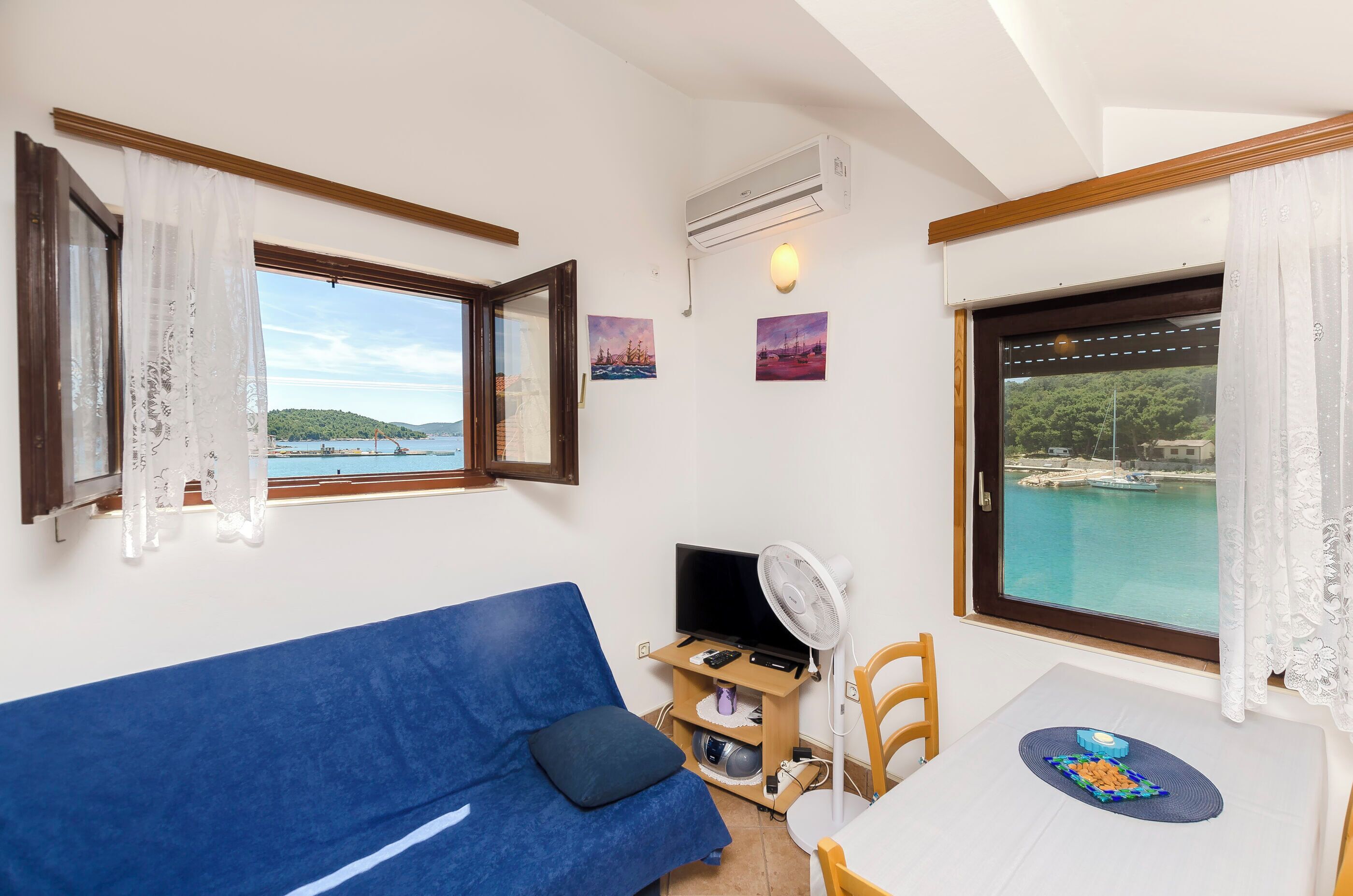 A room with a blue sofa, a dining table with a plate, a TV on a wooden stand, and a view of a body of water through a window.