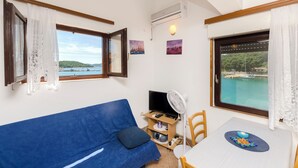 Una habitación con un sofá azul, una mesa de comedor con un plato, un televisor sobre un soporte de madera y vistas a un cuerpo de agua a través de una ventana.