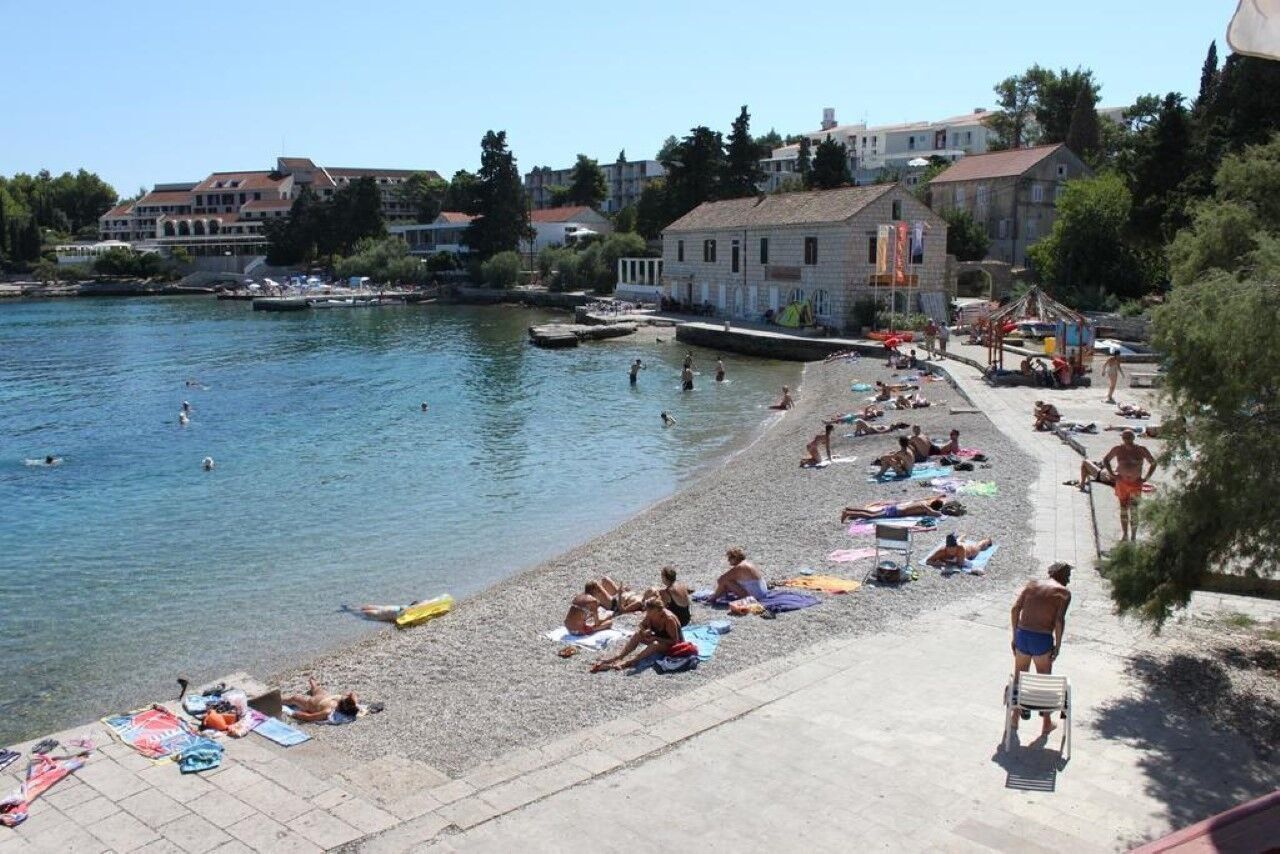 A beach with people sunbathing and swimming, buildings in the background, and a clear sky.
