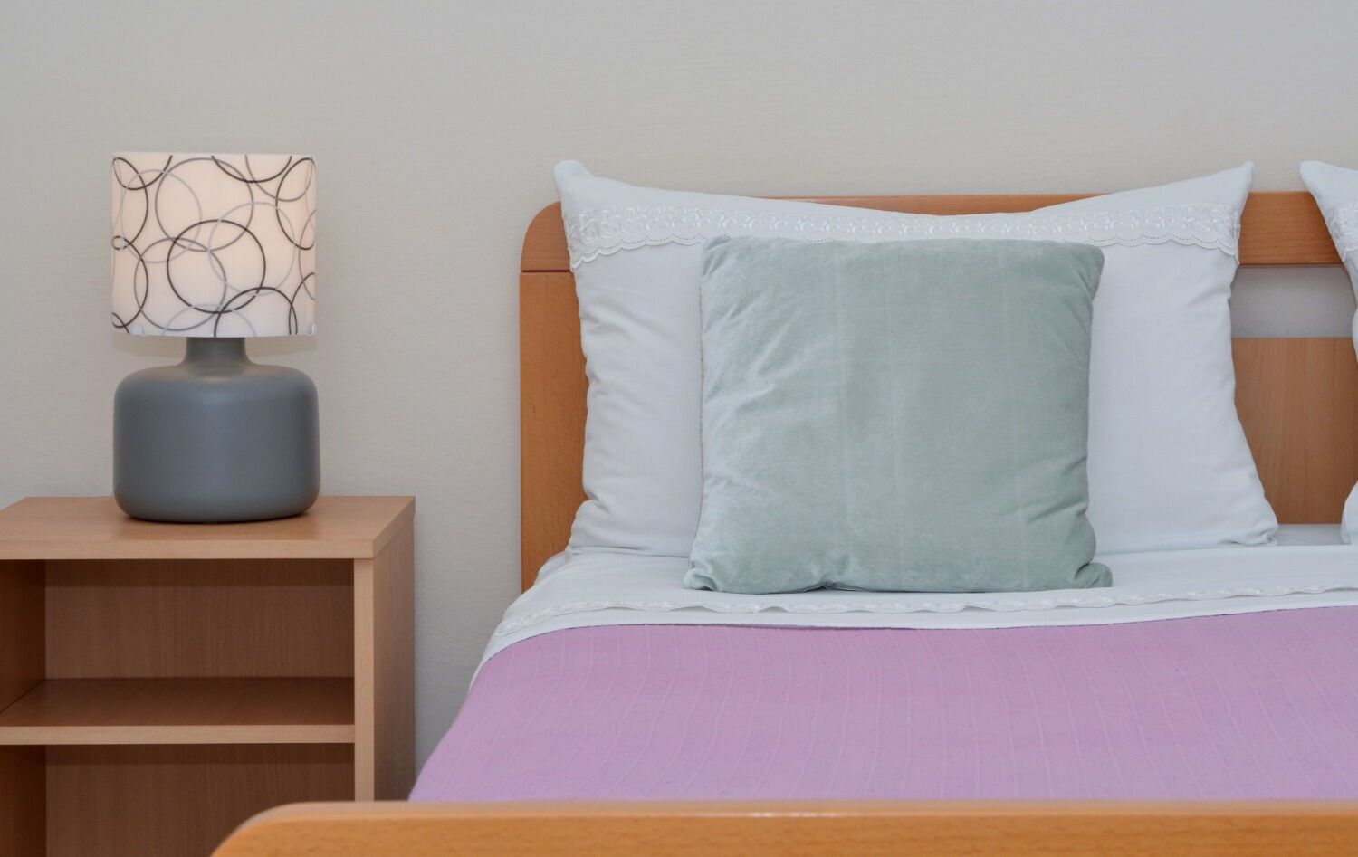 Ein ordentlich bezogenes Bett mit einem Holz-Kopfteil, ein Nachttisch mit Lampe und eine Wand mit einer geometrischen Leuchte.