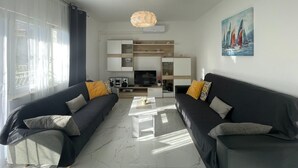 A modern living room with two dark sofas, a white coffee table, and a TV. There is a painting of sailboats on the wall, and the room has a large window with curtains. - Apartments Paz (Marina)