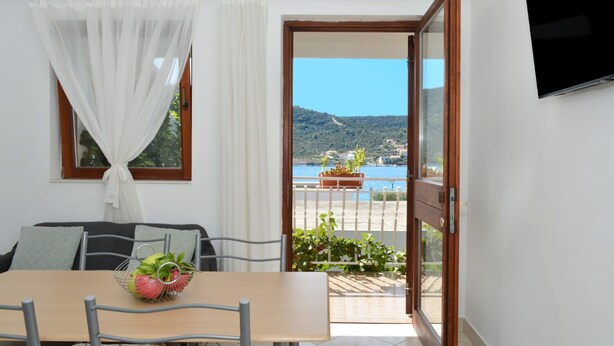 A room with a dining table, chairs, a sofa, and a view of a coastal landscape through an open door. - Apartments Paz (Marina)