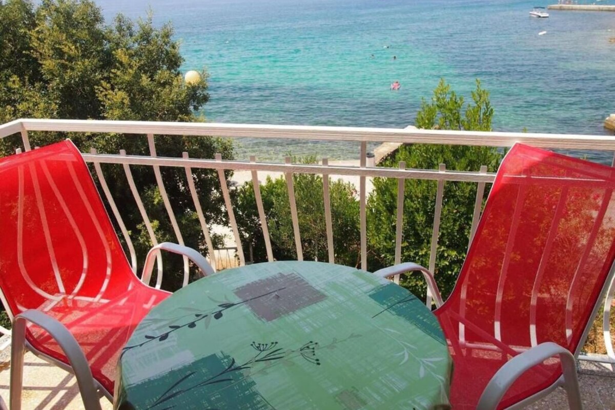 A balcony with two red chairs and a table with a green cushion, overlooking a clear sea with boats and a distant island.