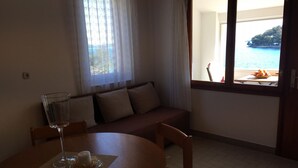 A dining area with a round table, a glass of water, and a dessert on a placemat. There is a sofa with cushions and a window with a view of the sea. - Apartments Durda (Sali)