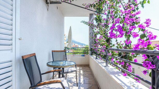 A balcony with a table and chairs, a railing, and purple flowers. - Apartments Josip (Baska Voda)