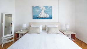 A bedroom with a large bed, two bedside tables with lamps, a painting of sailboats, a mirror, and an air conditioning unit. - Apartments Josip (Baska Voda)