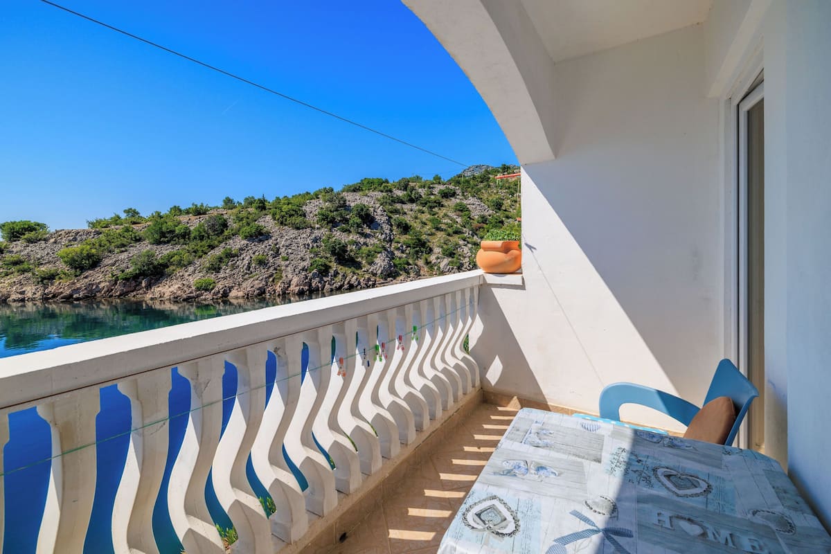 Un balcón con vista a un promontorio rocoso y una extensión de agua. Hay una mesa con mantel estampado y dos sillas.