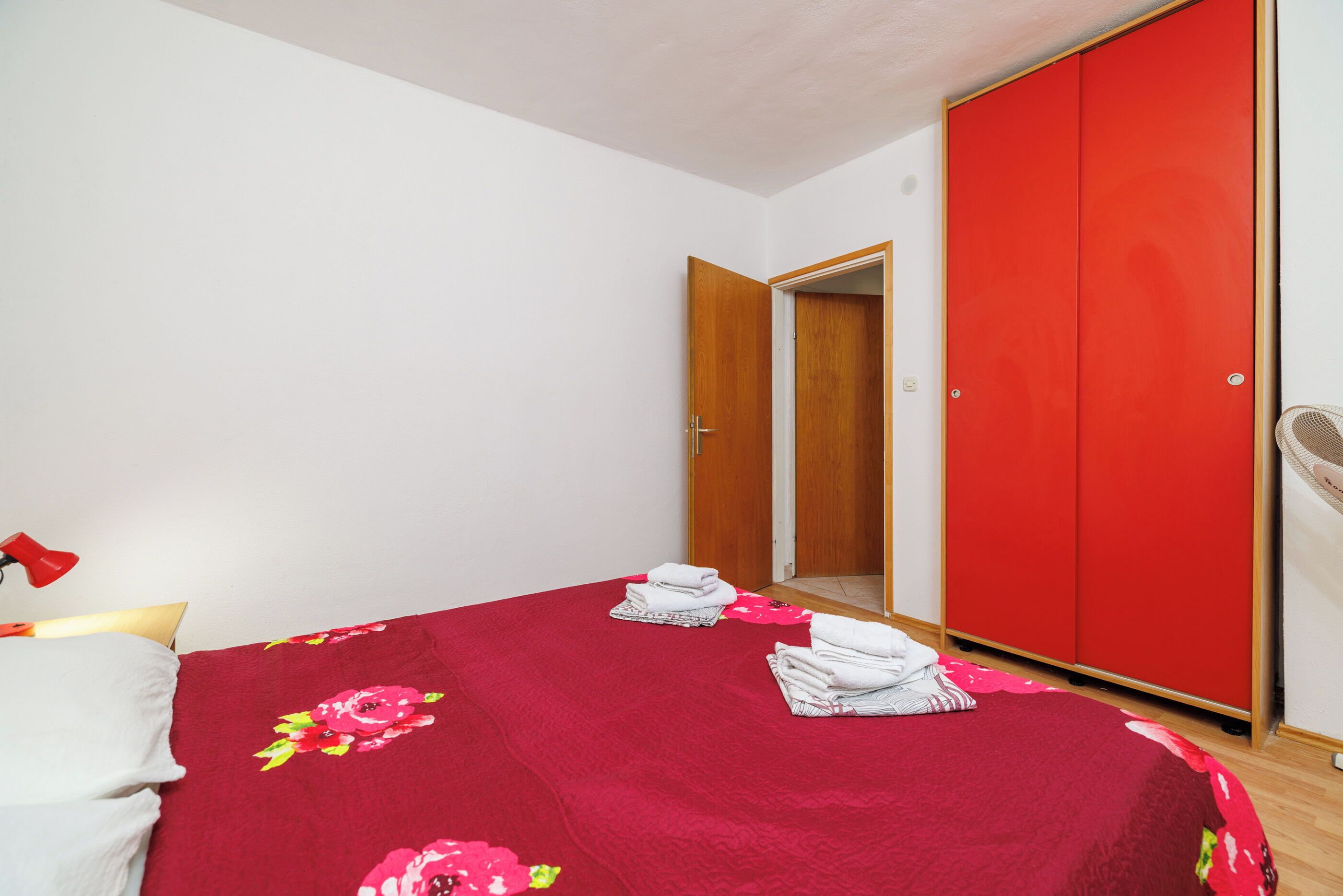 A bedroom with a red bedspread, a wooden wardrobe, and a white bedside table.