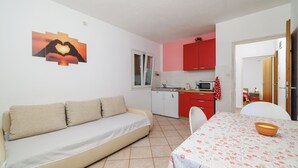 Un espace de vie compact comprenant un canapé, une table à manger recouverte d’une nappe à motifs floraux, un coin cuisine avec des placards rouges et une fenêtre agrémentée de rideaux.