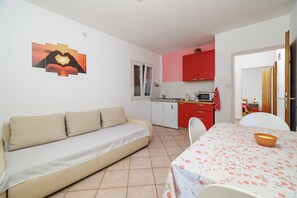 Ein gemütlicher Wohnraum mit einer Couch, einem Esstisch mit Blumenmuster und einer Küchenzeile mit roten Schränken, sowie einem Fenster mit Vorhängen.