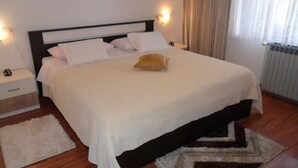 Een keurig opgemaakt bed met witte lakens en één decoratief kussen, een nachtkastje met een lamp en een raam met gordijnen.