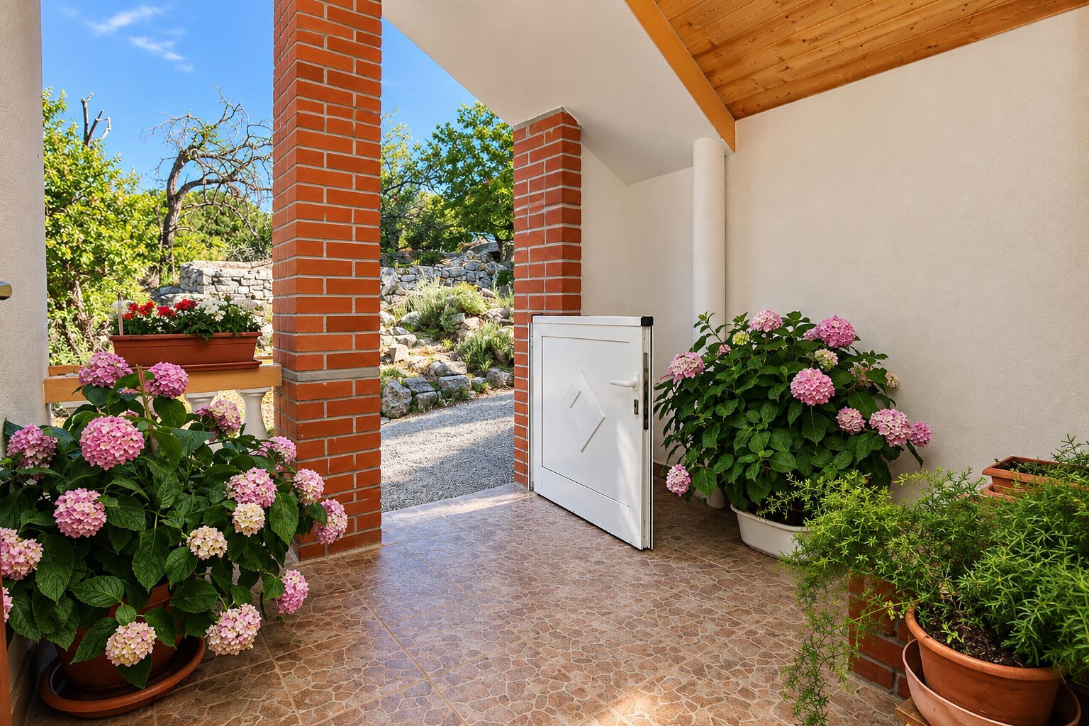 Un espacio al aire libre cubierto con un muro de ladrillo, una puerta blanca y macetas con flores rosadas.